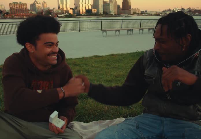 Two men smiling and greeting each other while sitting on grass by the riverside