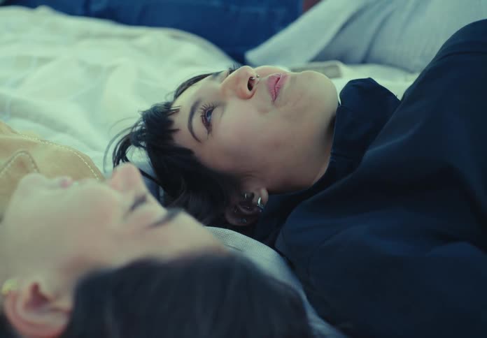 A close view of a smiling woman with dark hair and a nose ring wearing a dark shirt as she lies on a white blanket next to another person with soft natural lighting