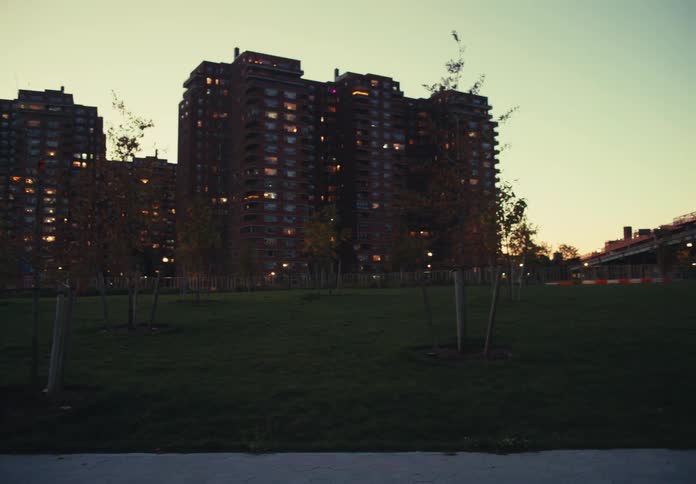 A lowangle camera pans right across a wide expanse of grass and small trees at dusk revealing dark tall apartment buildings with glowing windows on the left and a massive steel bridge structure on the right