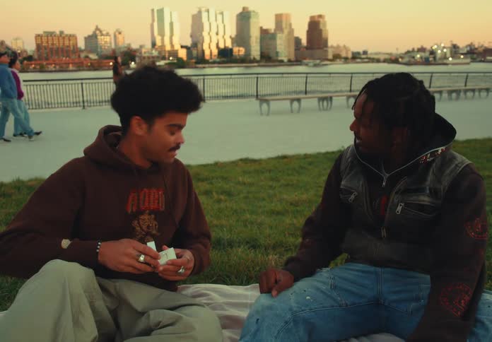 Two men sit facing each other on a blanket on a grassy lawn with an urban skyline backdrop where one man opens a ring box and presents it the other man approves it and then the first man stands up and walks away