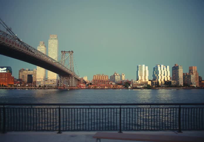 A bridge and a city skyline across the river are framed by a low metal railing in the foreground while people run and walk along the waterfront promenade under the fading light of dusk