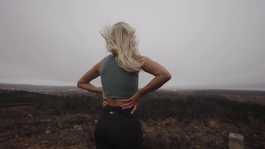 A woman with light hair wearing a tank top and dark pants stands with hands on her lower back on a dirt hilltop overlooking a vast landscape the wind blows her hair all under an overcast sky