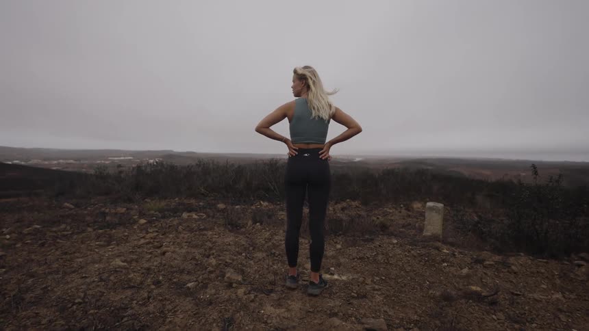 A wide shot from behind shows a blonde woman in a grey crop top and black leggings standing on a dark dirt hill with her hands on her hips looking out over a hazy landscape and the ocean on an overcast day