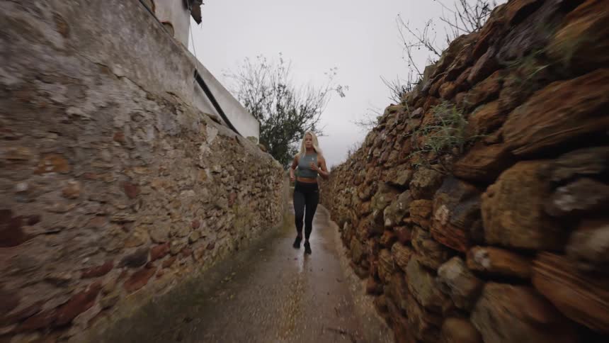 A low handheld view shows a blonde woman wearing a grey tank top and black leggings jogging through a narrow wet stone alleyway with rough stone walls under an overcast sky