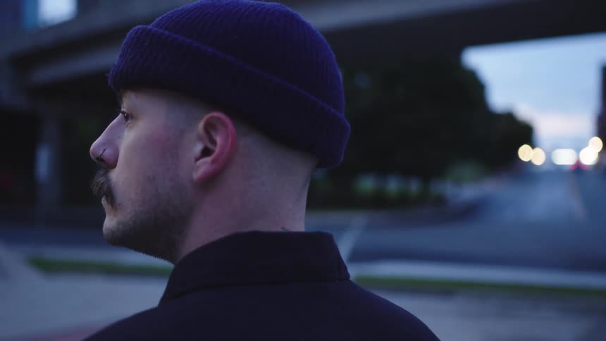 A man wearing a dark knit hat and dark jacket stands outdoors initially facing the camera in profile with a mustache and nose ring then turns to face away as car lights appear in the street behind him at dusk