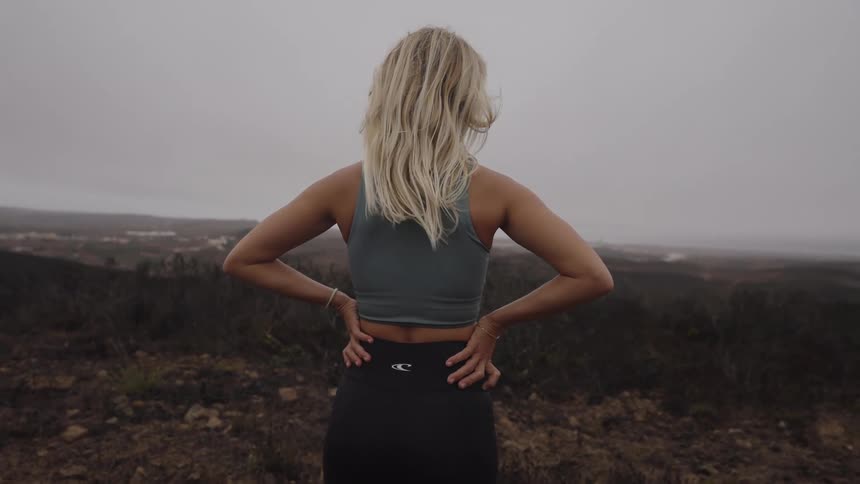 A woman with long blonde hair wearing a crop top and dark leggings stands on a hilltop with her back to the camera and her hands resting on her lower back looking out over a desolate landscape obscured by fog