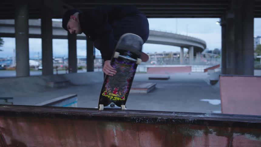 A person wearing a beanie skateboards under an overpass with city infrastructure visible beyond