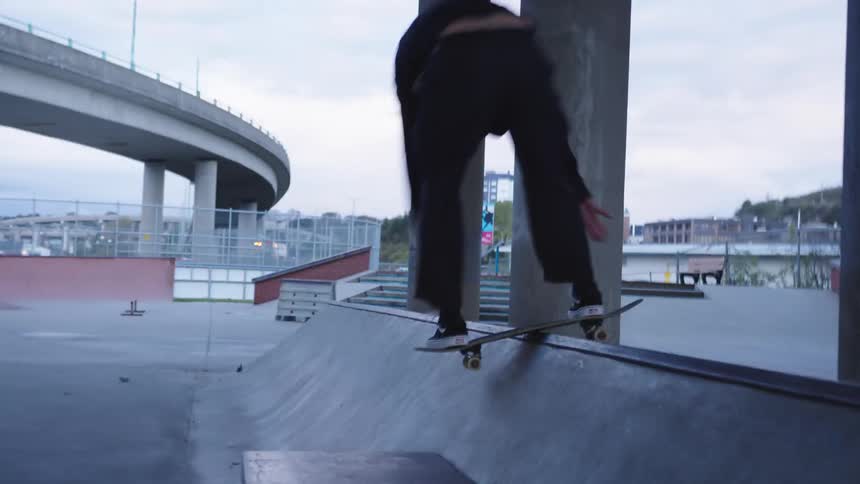 A person wearing dark clothes rides a skateboard slides along the edge of a ledge the skateboarder dismounts and lands on the ground in a skate park near an elevated road structure