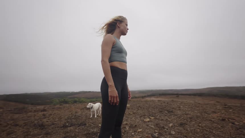 Woman in athletic wear gazes at the horizon amid a barren landscape with a white dog in the background