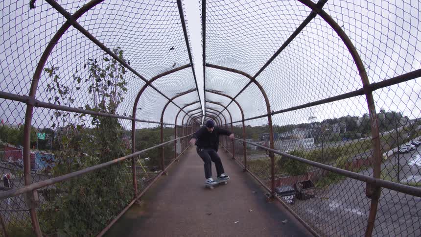 A person on a skateboard rides through a narrow caged walkway with a metal chainlink roof and walls connecting to an outdoor area