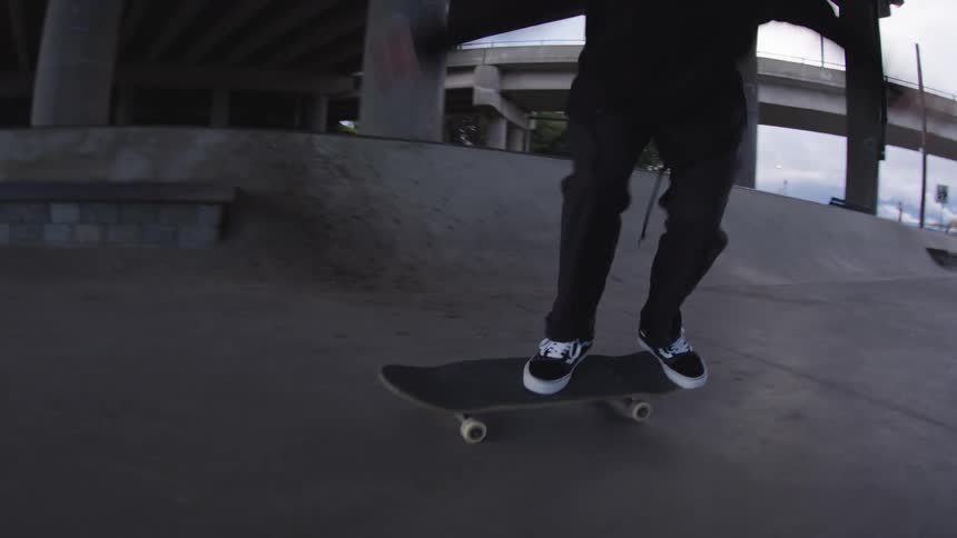 A person stands on a skateboard performs a trick across a concrete surface the setting is a skate park with ramps and pillars and a roadway structure is visible overhead