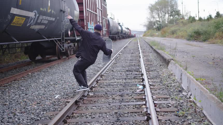 A person in dark clothing walks along a set of railway tracks balancing on one steel rail while holding a black skateboard and extending an arm for balance with long freight train cars stopped on an adjacent track