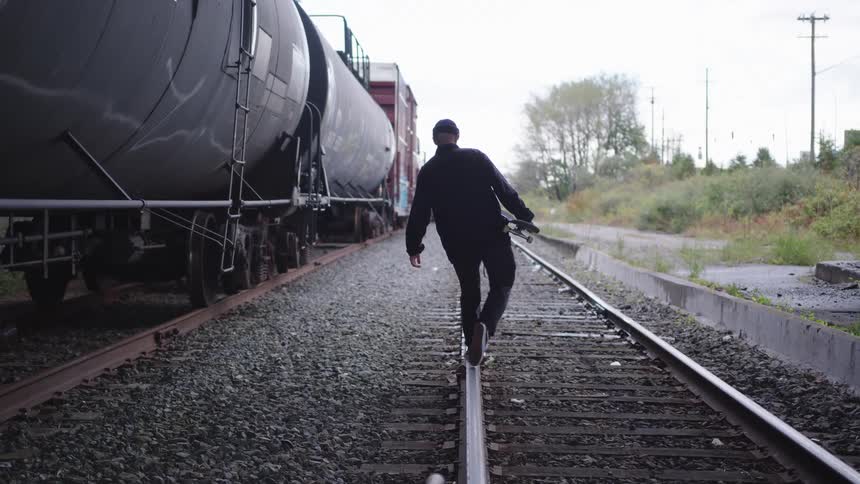 Man walks along rail tracks beside freight cars holding a skateboard