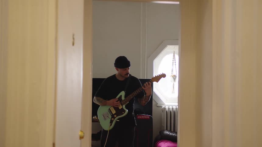 The video is framed through a doorway showing a person standing in a brightly lit room playing a light green electric guitar they are wearing a dark beanie and shirt and appear to be practicing or performing privately
