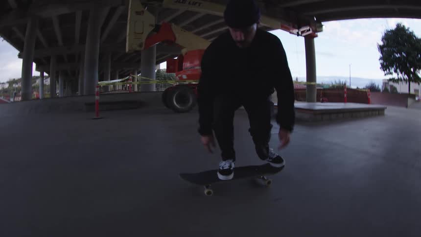 A man wearing all black clothing and a beanie skateboards in a concrete skate park under a freeway overpass attempting a trick before falling to the ground