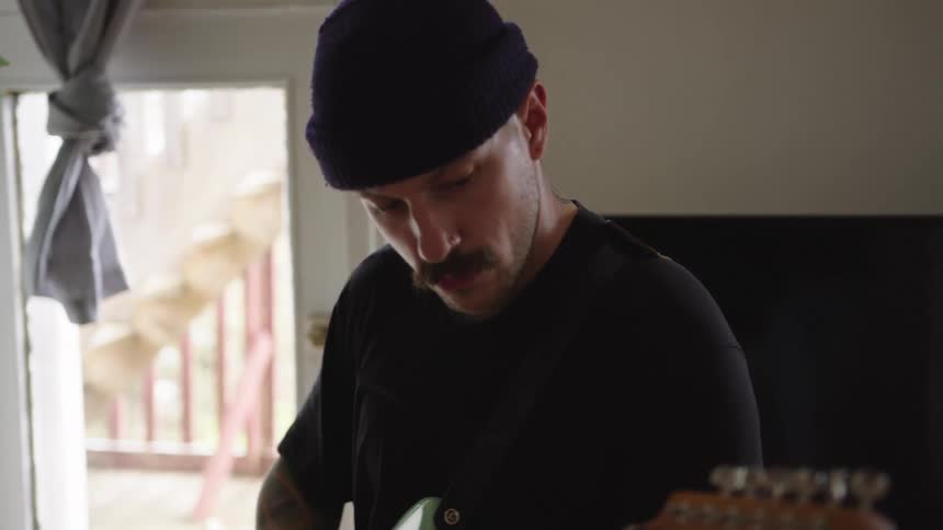 A man wearing a dark beanie and a mustache looks down in concentration while playing an electric guitar indoors near a bright glass door
