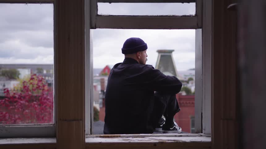 A man in a purple beanie sits on a window ledge overlooking the city appearing quiet and reflective
