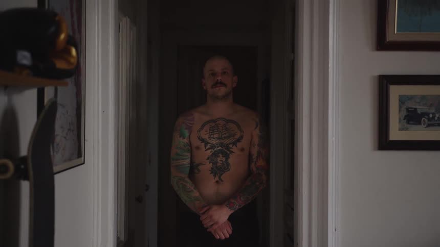 A shirtless man with many tattoos stands in a residential home