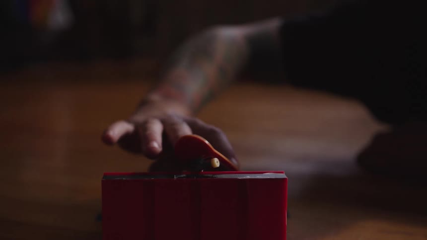 A person with a tattooed arm plays with a toy skateboard on a wooden floor indoors at midday sunlight filtering through the windows and casting soft shadows across the room