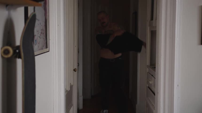 Man removes his shirt in a narrow hallway with a skateboard leaning against the wall