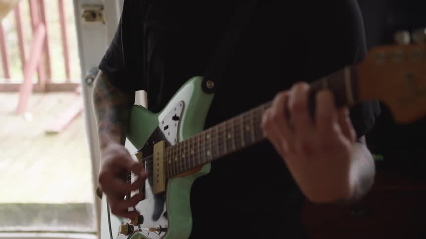 White man wearing dark clothes strumming on an electric guitar next to a window indoors during day time