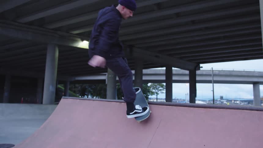 A low angle dynamic view shows a man wearing a dark jacket pants and a knit cap skateboarding in a skatepark doing a trick on the lip of a ramp under a freeway overpass on a dull day