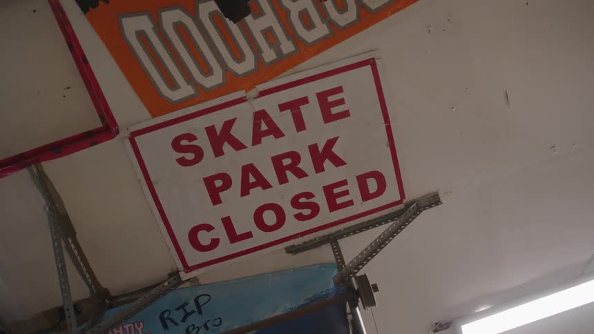 A skateboarding park sits empty under the midday sun with a sign posted at the entrance that reads skate park closed