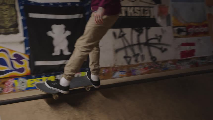 A person in tan pants rides a skateboard performing a grind trick along a dark narrow rail in the foreground of a skatepark with graffiticovered walls