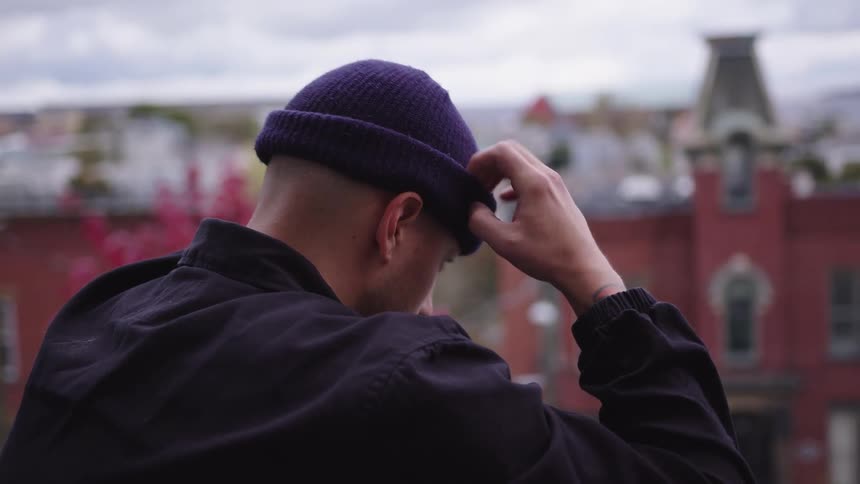Man wearing a beanie looks out over the city while leaning on a balcony