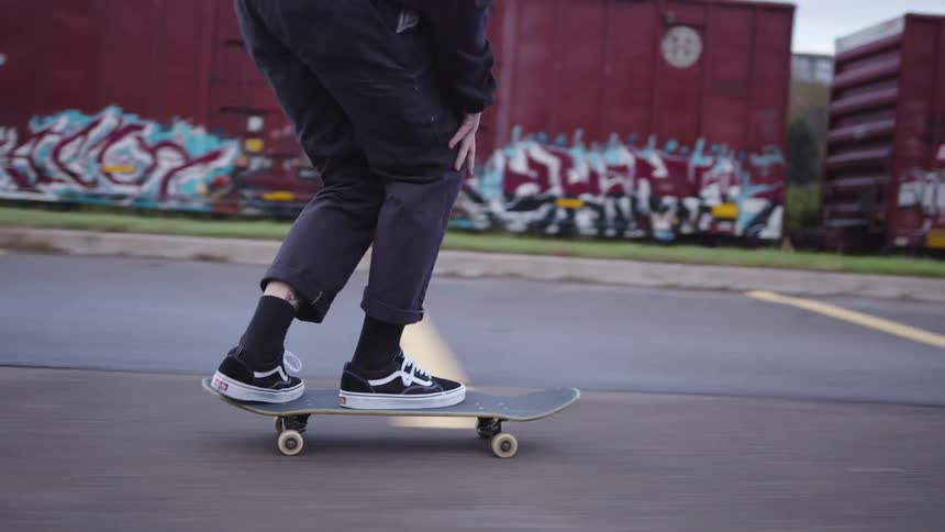 A person wearing dark pants and black and white shoes pushes a skateboard and glides on a paved surface with yellow markings moving past large red cargo containers or train cars and oncoming automobiles