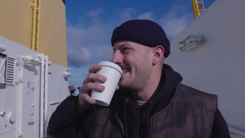 A young man wearing a jacket and hat walks outdoors at midday smiling as he drinks a cup of coffee
