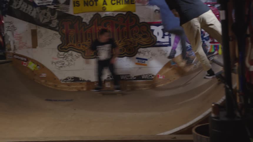 The video is an interior shot of an indoor skatepark filled with bright graffiti and posters including a sign that reads skateboarding is not a crime several young men are skateboarding and practicing tricks on a wooden halfpipe ramp