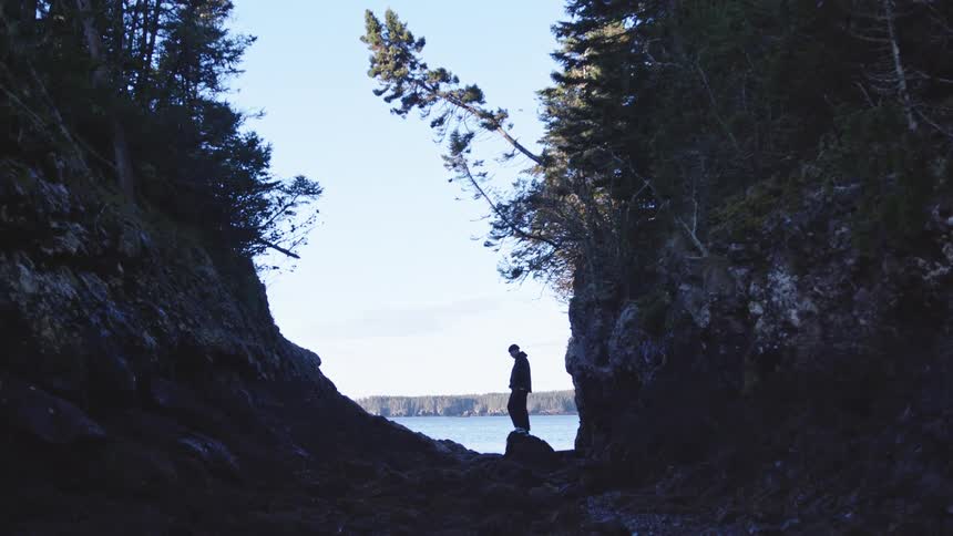 A male figure stands on a rocky shoreline with a lake view in the background at midday