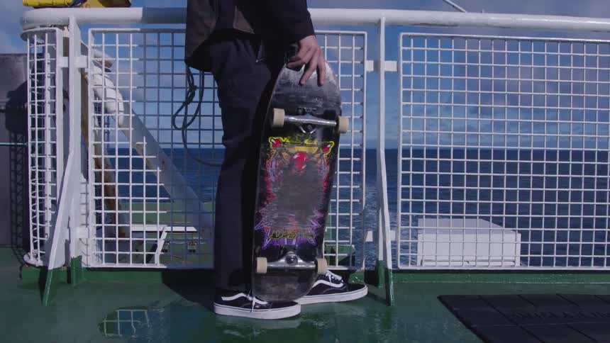 Person holds a skateboard resting on the floor while looking at the sea on the deck