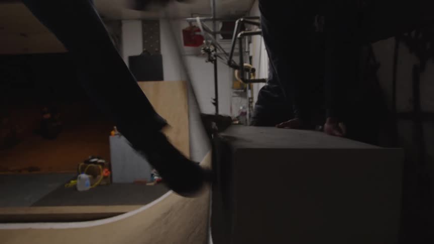 The video shows a closeup of a person skateboarding on a small smooth wooden ramp inside a dark garagelike space the walls around the ramp are covered with an assortment of posters and stickers suggesting a makeshift indoor skate spot