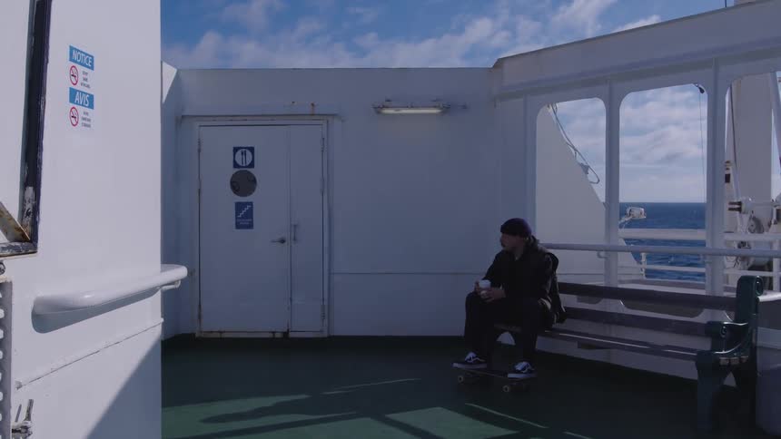 A man wearing dark clothing and a cap sitting on a wooden bench on the outdoor deck of a ship drinking from a cup while his skateboard rests under his feet with the open water visible beyond the railing