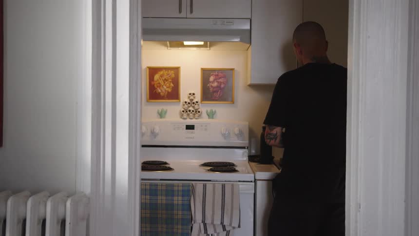 The video is a static shot of a small kitchen seen through a doorway with a man standing at the counter in the right foreground a white stove is visible and above it are two framed pictures and a decorative sculpture of skulls