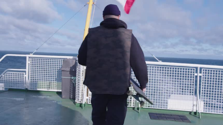 A person wearing a dark hooded sweatshirt and a dark vest walks away from the camera on the open deck of a moving vessel carrying a skateboard toward a white railing overlooking the blue water