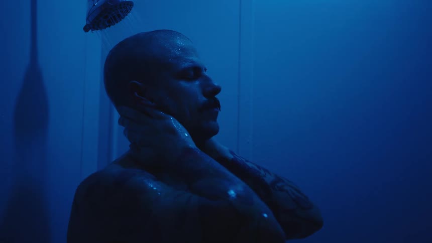 A man stands in a shower illuminated by a blue light washing his head and face with his hands under the stream of water