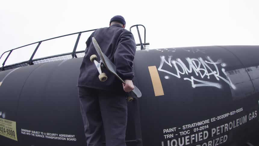 Man holds a skateboard while climbing the side ladder of a train car
