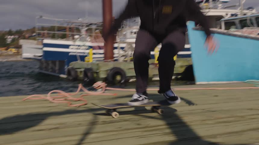 The video shows a man skateboarding on a wooden dock with several boats including a prominent blue fishing vessel tied up behind him he attempts a trick bringing the nose of the board up before the short clip ends