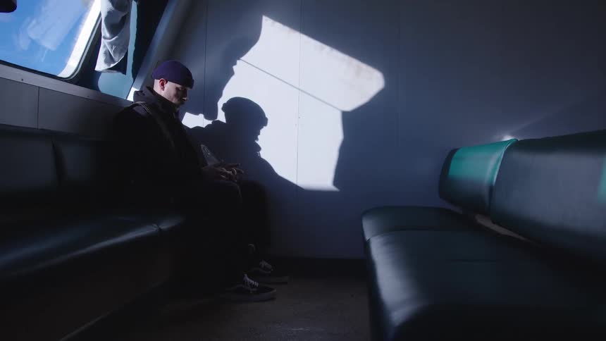 A moody interior view shows a man wearing a dark knit cap and jacket sitting alone on a dark bench inside a moving train or ferry illuminated by a bright shape of sunlight casting a shadow on the white wall