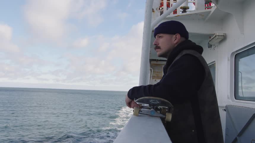 A man with a mustache wearing a black cap and jacket stands on the deck of a white ship holding a skateboard and looking out over the ocean under a cloudy sky