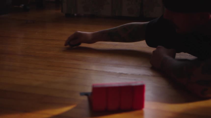 A person kneels on a wooden floor and uses their fingers to operate a small fingerboard toy the tiny skateboard rolls and flips off the floor and a red rectangular object sits in the blurred foreground