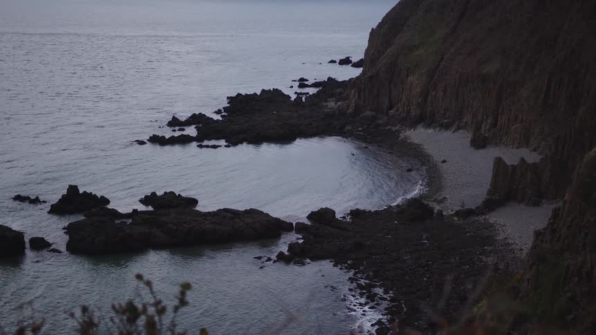The video provides an elevated view of a rocky dark gray cove and shoreline where the land meets the water the dark rocks create a semicircle around a small patch of dark sand or shingle beach