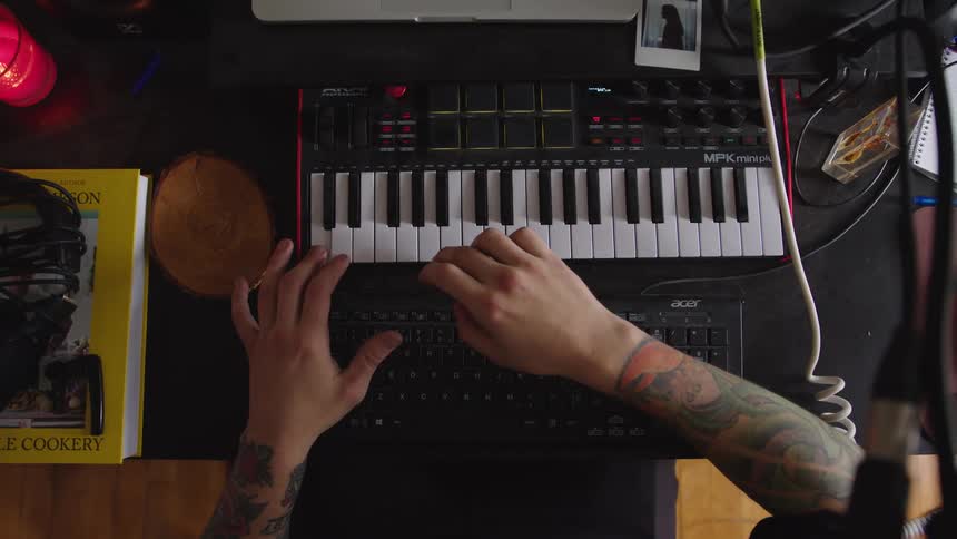 An overhead view shows the tattooed arms and hands of a person playing a black and white midi keyboard controller placed on a dark desk next to a computer keyboard a cookbook and other sound equipment