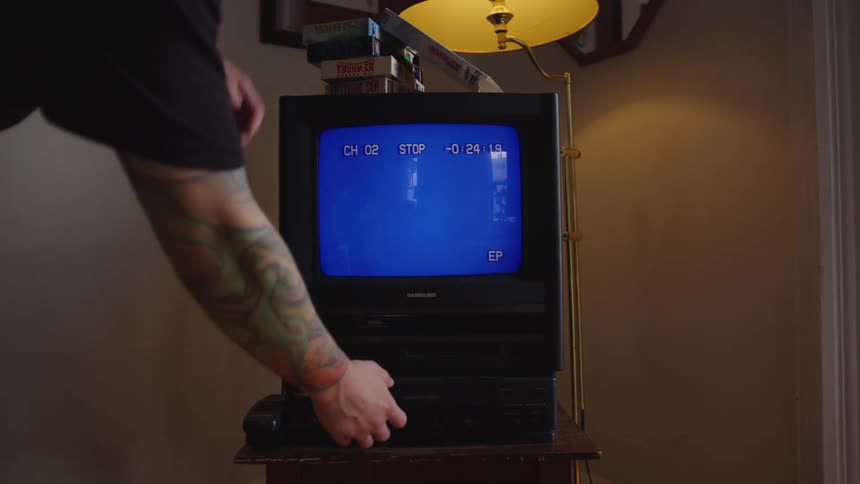 A person stands over a television and vcr the television shows skateboarding footage the person reaches down and stops the vcr the screen turns blue the person removes the videocassette and puts it on the case