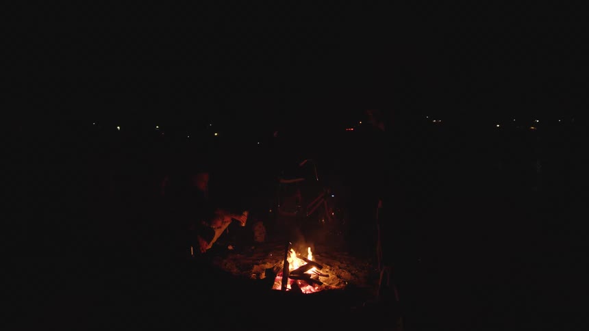 A bright fire burns in a small outdoor pit providing the only light for a group of people whose silhouettes are visible around the flames against a very dark background with faint distant lights
