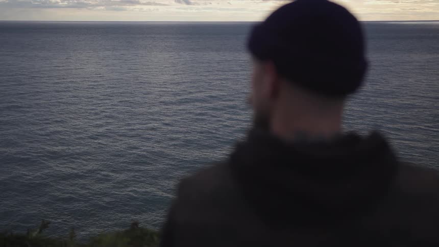A person wearing a dark beanie and dark jacket stands with their back to the camera looking out over a wide expanse of deep gray rippling water under a light gray sky with a visible horizon