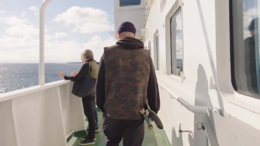 A man wearing a dark knit cap and a camouflage vest walks away from the camera on the white deck of a ferry or large boat carrying a skateboard as other passengers look out over the blue ocean on a sunny cloudy day